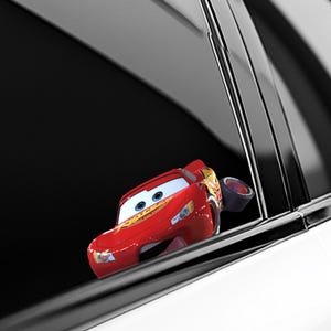 May include: A red Lightning McQueen car-shaped accessory, featuring blue eyes and a smiling face, is attached to a car window. The accessory has a black tire attached to the side. The car window is black and the car is white.
