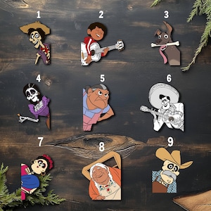 May include: A collection of nine colorful wooden cutouts featuring characters from the animated movie "Coco." The characters include Miguel playing guitar, a skeleton with a hat, a dog with a bone, and other figures in various poses. The cutouts are set against a dark wood background.