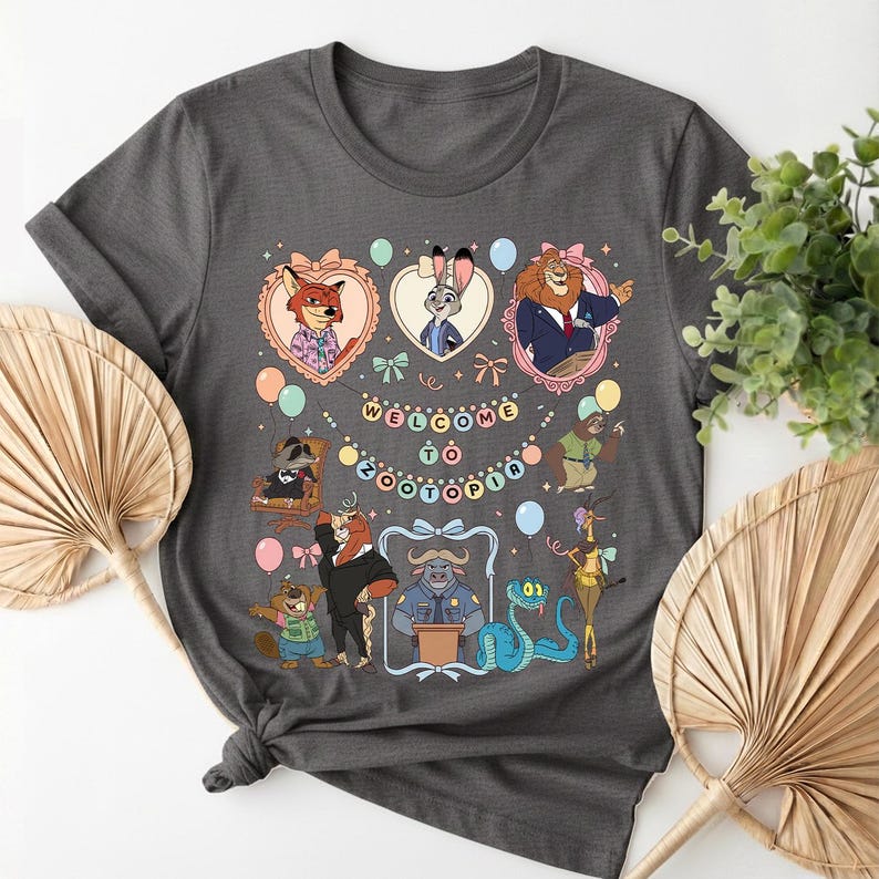 K&ouml;nnte beinhalten: Dunkelgraues T-Shirt mit einem farbenfrohen Grafikdesign von Zootopia-Charakteren. Das Design umfasst gerahmte Portr&auml;ts eines Fuchses, Kaninchens und L&ouml;wen sowie den Text "WELCOME TO ZOOTOPIA" und andere Zeichentrickfiguren.