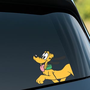 May include: A yellow Pluto cartoon character sticker with a green collar and pink tongue, running to the right. The sticker is applied to a car window, with a dark background and a glimpse of the outdoors.