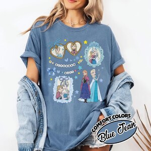 May include: A blue t-shirt featuring a graphic print of two animated characters in heart-shaped and ornate frames, along with the text "birthday girl" on a string of beads. The design includes blue bows, stars, and snowflakes on a solid blue fabric.