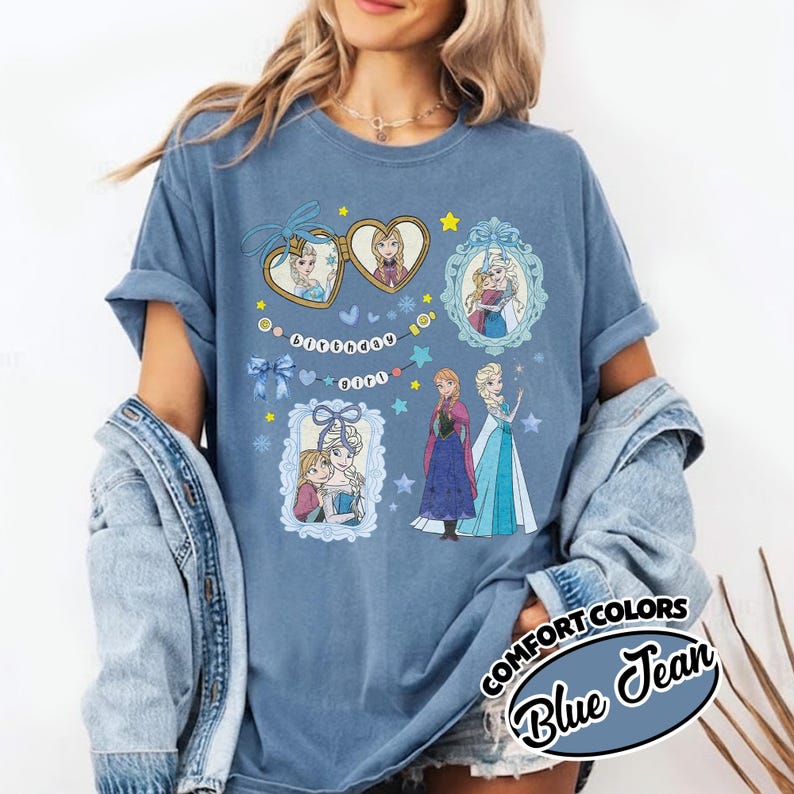 May include: A blue t-shirt featuring a graphic print of two animated characters in heart-shaped and ornate frames, along with the text "birthday girl" on a string of beads. The design includes blue bows, stars, and snowflakes on a solid blue fabric.