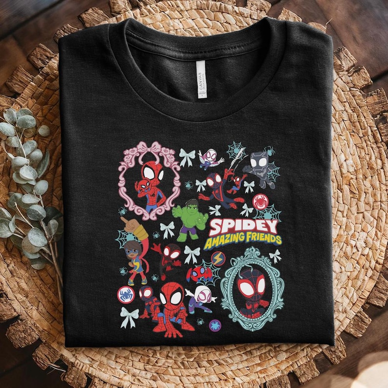 May include: Black t-shirt featuring a colorful graphic of Spider-Man and other characters. The design includes the text "Spidey and His Amazing Friends" with decorative frames, bows, and spiderwebs. The shirt is folded on a woven surface.