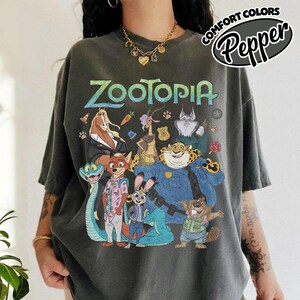 May include: A charcoal gray short-sleeve t-shirt featuring a large graphic print of Zootopia characters including Judy Hopps, Nick Wilde, and Chief Bogo. The text Zootopia is printed in large teal letters above the colorful character illustration.