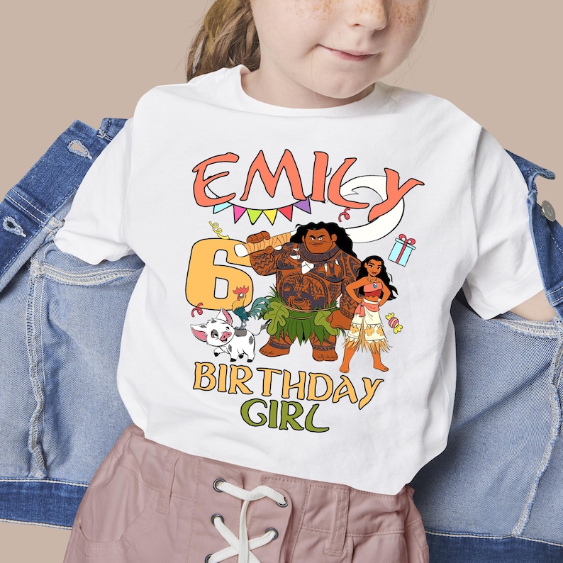 May include: A white t-shirt featuring a graphic print with the name EMILY in orange, the number 6 in yellow, and the text BIRTHDAY GIRL in green. The design includes cartoon characters, a pig, a gift box, and colorful bunting flags.