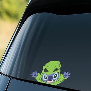 May include: A colorful vinyl decal featuring a cartoon character, Stitch, in a green costume. The character has blue skin, large eyes, and is smiling. The decal is applied to a car window.