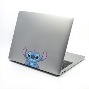 May include: A silver laptop with a cartoon sticker of Stitch, a blue alien character with large ears and big eyes. The sticker is placed on the lower part of the laptop's lid. The laptop is on a white surface.