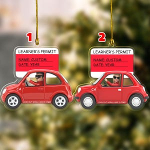 May include: Two hanging ornaments shaped like red cars with white wheels. Each features a white and red sign reading LEARNER'S PERMIT, NAME: CUSTOM, DATE: YEAR, and the text LOOK OUT WORLD HERE I COME! One car contains a figure with sunglasses, the other a figure with blonde hair and sunglasses.