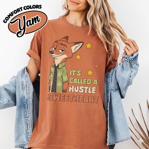 Puede incluir: Una camiseta de manga corta color &oacute;xido con el gr&aacute;fico de un zorro de dibujos animados con camisa verde y corbata. El texto dice "IT'S CALLED A HUSTLE SWEETHEART" junto a estrellas amarillas. La prenda se lleva bajo una chaqueta vaquera azul claro.