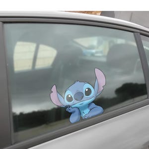 May include: A blue and purple cartoon character sticker of Stitch from Lilo & Stitch, applied to a car window. The character is in a relaxed pose, with one hand under its chin.