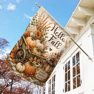 May include: A rectangular garden flag featuring a wooden wheelbarrow filled with pumpkins, wheat, and sunflowers. The text reads Hello Fall with a small heart, and a wooden sign below says Grateful Thankful Blessed. The background is a light cream color.