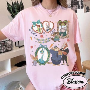 May include: A light pink t-shirt featuring a colorful graphic print of various animated animal characters from Zootopia, including a fox, rabbit, snake, and cheetah, framed by decorative borders and the text Welcome to Zootopia in a banner.