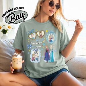 May include: A sage green short-sleeve t-shirt featuring a graphic print of two cartoon characters in heart-shaped and rectangular frames, along with the text "Birthday Girl" on a banner. The design includes blue bows, stars, and snowflakes.