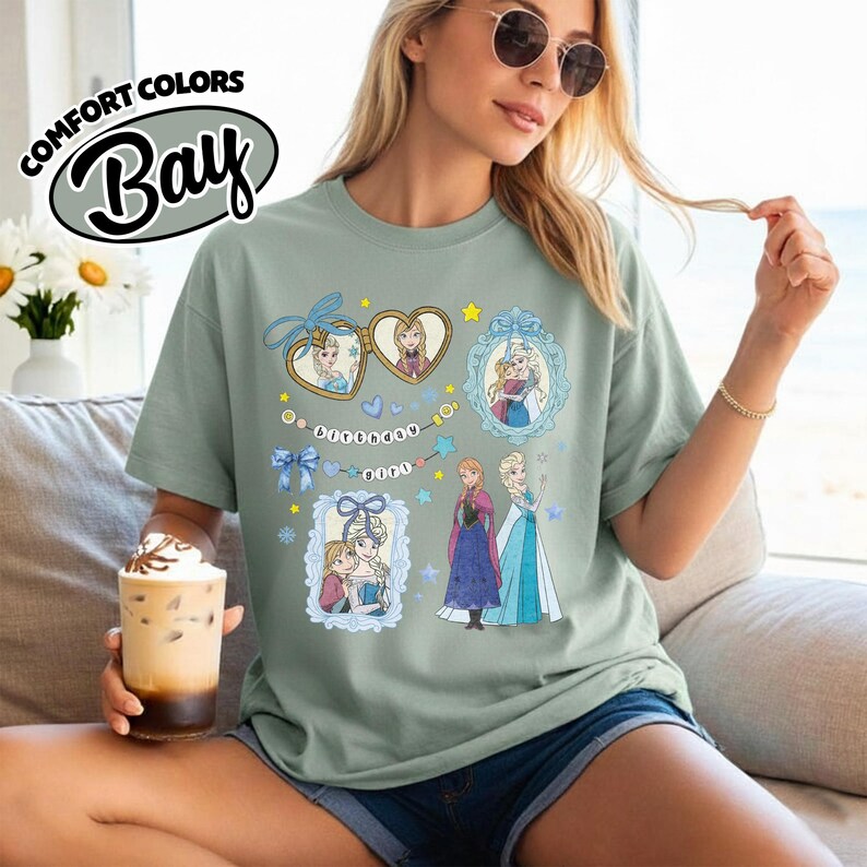 May include: A sage green short-sleeve t-shirt featuring a graphic print of two cartoon characters in heart-shaped and rectangular frames, along with the text "Birthday Girl" on a banner. The design includes blue bows, stars, and snowflakes.