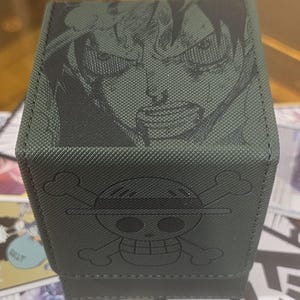 May include: Green card storage box featuring a detailed illustration of a character's face on the top and a skull and crossbones emblem on the front. The box is designed to hold trading cards and has a textured surface.