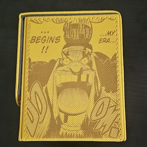 May include: Yellow wallet with a brown manga-style illustration and text. The wallet features the phrases "BEGINS!!" and "...MY ERA..." in a comic book font. The design includes a character with an open mouth and the sound effect "DOOOW!"
