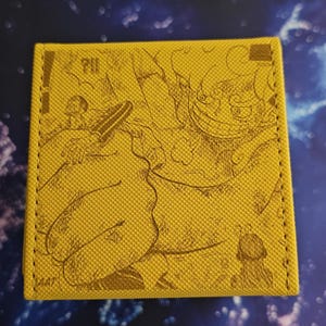 May include: Yellow wallet with a textured surface and a manga-style illustration in brown. The wallet features a stitched border and a detailed drawing of a character. The text "?!!" is visible.