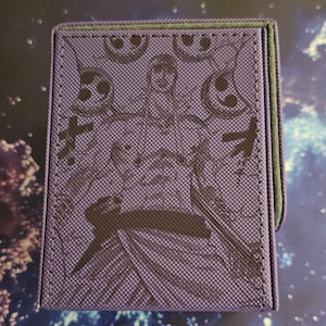 May include: A square, purple card case with a textured surface. The case features a detailed illustration of a character with Japanese text. The edges are trimmed with green stitching.