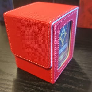 May include: A red, rectangular card storage box with white stitching and a clear window. The box has a hinged lid and is designed to hold trading cards. Other boxes are visible in the background.