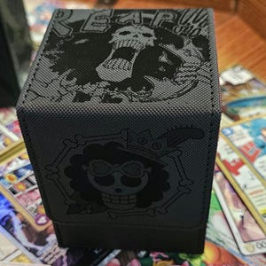 May include: A black card storage box featuring a skull design with the text "REAP". The box has a textured surface and a hinged lid. The box is surrounded by colorful trading cards.