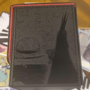 May include: Black card storage box with a red trim. The box features a textured surface and a black silhouette design. The box is designed to hold trading cards.