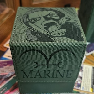 May include: Green box with a black illustration of a character's face on the top. The front of the box has the word "MARINE" in black, with a symbol above it. The box is sitting on a wooden surface.