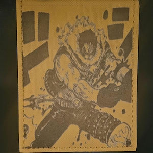 May include: A tan fabric patch with a black and white image of a character from a manga or anime. The character is wearing a fur-lined coat and has a large, spiky hairstyle. The patch has a stitched border.
