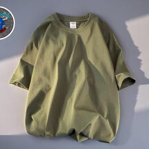 Op de afbeelding: Olijfgroen T-shirt met korte mouwen en een ronde hals. Het shirt is gemaakt van een zachte, lichte stof. Een cirkelvormige afbeelding met tekst en een blauw ontwerp is zichtbaar linksboven.