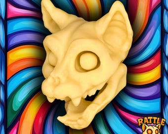 May include: A light beige cat skull with open mouth and visible teeth. The skull is set against a vibrant, swirling background of rainbow colors. The image is framed with a blue border and includes the text "Rattle Pets".