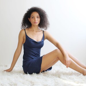 May include: A woman wearing a navy blue slip dress with a cowl neck sits on a white fluffy rug.