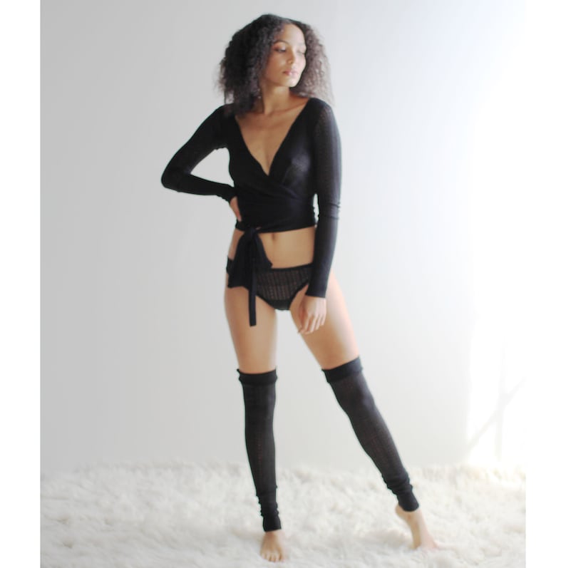 May include: A woman wearing a black long-sleeved wrap top, black lace underwear, and black leg warmers. She is standing on a white fluffy surface.