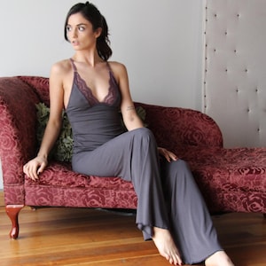 May include: A gray pajama set featuring a camisole top with a lace trim and wide-leg pants. The top has a halter neck design. The set is made of a soft, flowing fabric. The model is sitting on a red chaise lounge.