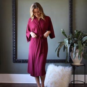 May include: A burgundy-colored robe with a wrap-around design and a matching tie belt. The robe has wide sleeves and falls to the mid-calf. The model is wearing the robe in a room with a large mirror and a potted plant.