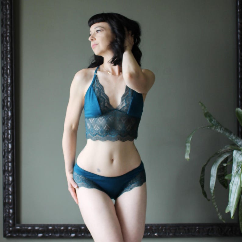 May include: Teal lingerie set featuring a bralette with a scalloped lace trim and matching low-rise briefs. The bralette has a halter neck and a delicate lace design on the cups. The set is made of a soft, comfortable fabric.