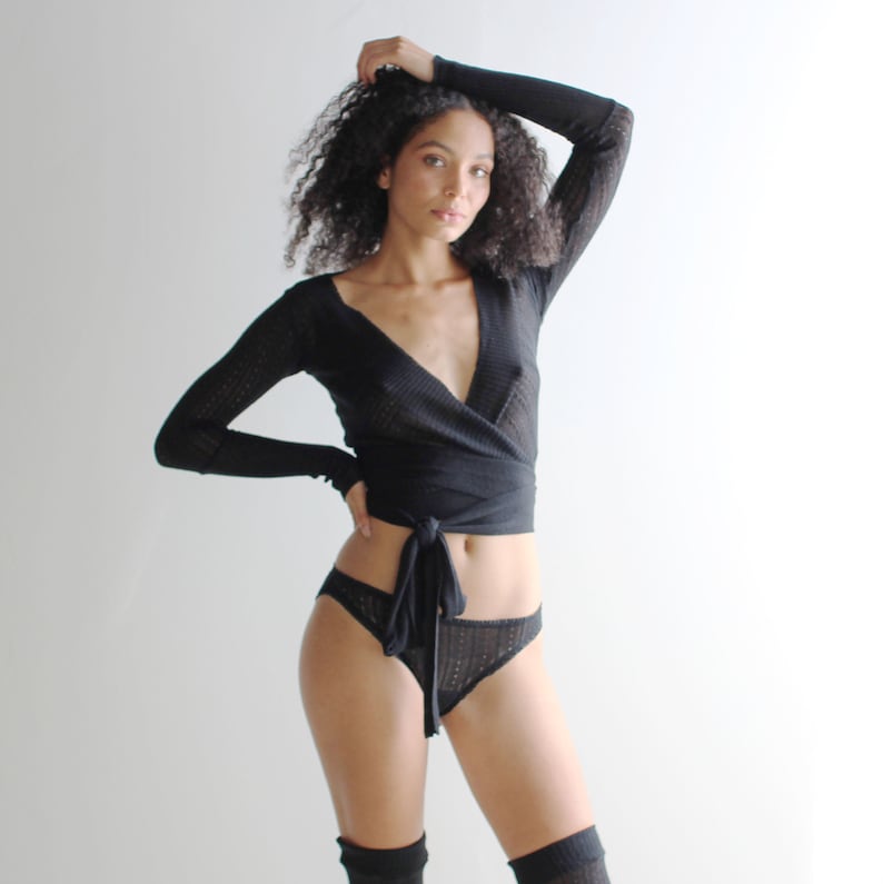 May include: A black wrap top with long sleeves and a deep V-neckline. The top is made of a soft, stretchy fabric and has a delicate knit pattern. The model is wearing the top with black underwear and black stockings.