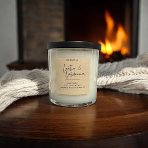 May include: A clear glass candle jar with a black lid, labeled "Leather & Labdanum." The label is a light brown rectangle with black text. The candle is 12 oz. and handmade in South Vienna, OH. The background shows a fireplace.