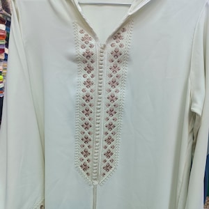 Peut inclure: Robe à capuche blanc cassé avec broderies florales complexes sur le devant et les manches. La robe a une coupe longue et ample et une fermeture éclair. La broderie est de couleur rose pâle.