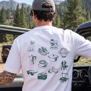 Könnte beinhalten: Ein weißes Kurzarm-T-Shirt mit einem Raster aus dunkelgrünen Strichzeichnungen auf der Rückseite. Die Grafiken zeigen einen Hund, Turnschuhe, eine Kaffeemaschine, einen Globus, einen Topf, eine Kaffeetasse, eine Sonnenbrille, eine Kamera, Berge, Tänzer und einen Plattenspieler.