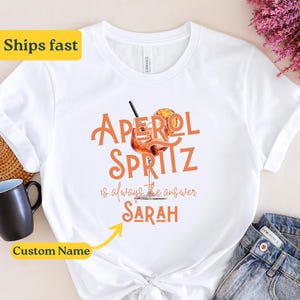 Könnte beinhalten: Weißes T-Shirt mit dem Text "Aperol Spritz is always the answer" und einem Glas Aperol Spritz. Der Name "SARAH" steht unter dem Text.