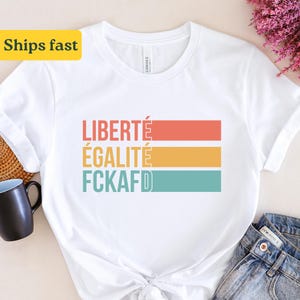 Może przedstawiać: Biała koszulka z retro grafiką przedstawiającą słowa "LIBERTÉ ÉGALITÉ FCKAFD" w kolorowym pasiastym wzorze.