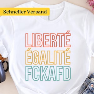 Könnte beinhalten: Ein weißes T-Shirt mit kurzen Ärmeln und einem dreizeiligen Grafikdruck. Der Text lautet LIBERTÉ in Koralle, ÉGALITÉ in Gelb und FCKAFD in Blaugrün. Die Schrift ist in einem fetten, serifenlosen und umrandeten Stil mittig platziert.