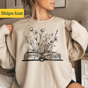 Könnte beinhalten: Beigefarbener Pullover mit einem schwarz-weißen Aufdruck eines geöffneten Buches mit Blumen und dem Text "One More Chapter".