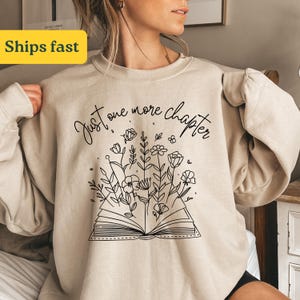Könnte beinhalten: Ein beiger Pullover mit dem Text "Just one more chapter" und einer schwarzen Strichzeichnung eines offenen Buches, aus dem Blumen wachsen.