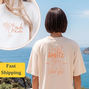 Puede incluir: Camiseta color crema con texto y gráficos en naranja. La parte delantera presenta "Bride Kathi" con una ilustración de cóctel. La parte trasera dice "One last Spritz" con la fecha "Junio 2026" y "(said yes!)" debajo de dos copas de cóctel.