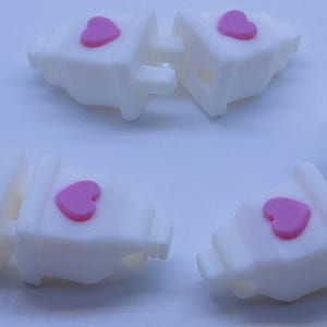 May include: Five white plastic plug-shaped charms with pink heart accents. The charms are designed to be used as zipper pulls or bag charms.