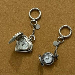 Peut inclure: Deux porte-clés de montre de poche argentés. L'un est en forme de cœur avec un couvercle ouvert, l'autre est rond avec un couvercle ouvert. Les deux ont des cadrans blancs avec des chiffres et des aiguilles noirs. Chaque porte-clés a un fermoir et un petit charme circulaire.