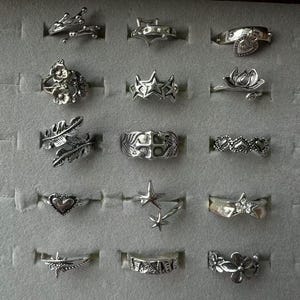 Könnte beinhalten: Eine Sammlung von fünfzehn silberfarbenen Metallringen, die in einem Raster angeordnet sind. Die Designs umfassen Blumenmotive, Sterne, Herzen, Federn, ein Kreuz und geometrische Muster. Jeder Ring weist feine Details und ein poliertes Finish auf.