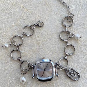 May include: A silver watch with a square face and a chain link bracelet. The bracelet has several charms, including a flower and pearls.