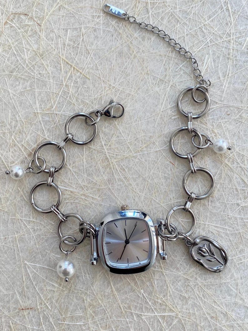 May include: A silver watch with a square face and a chain link bracelet. The bracelet has several charms, including a flower and pearls.