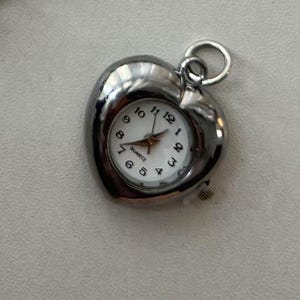 May include: A silver heart-shaped pendant watch with a white face and black numbers. The watch is quartz and has a small second hand.
